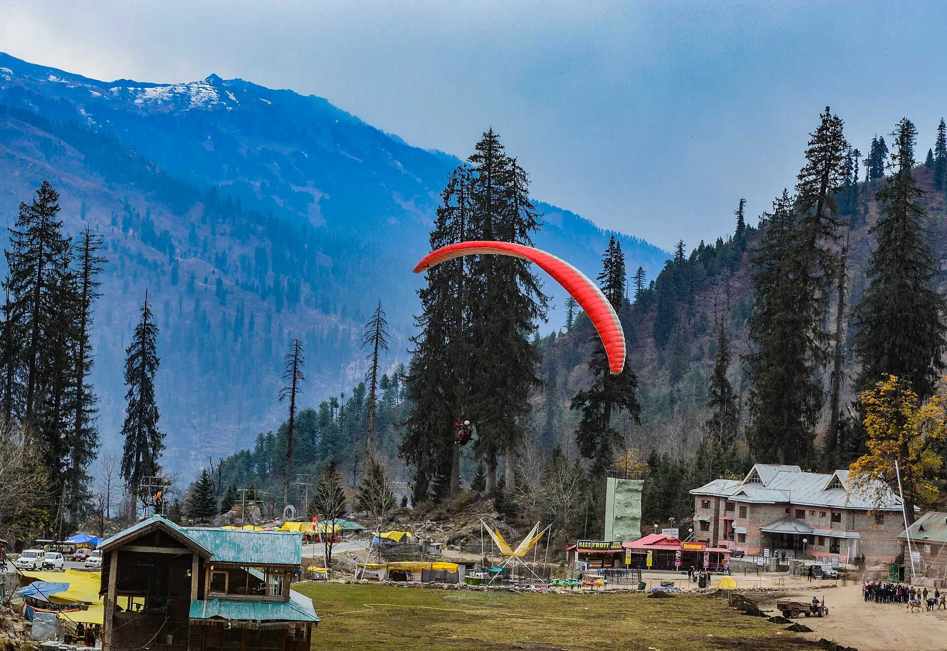 A paraglider's red canopy above pine trees and cabins in Solang Valley, with snowy mountains beyond